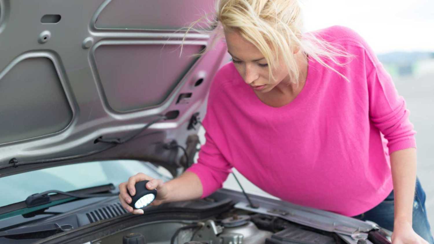 Self-sufficient confident modern young woman inspecting broken car engine with a flashlight in her hand.