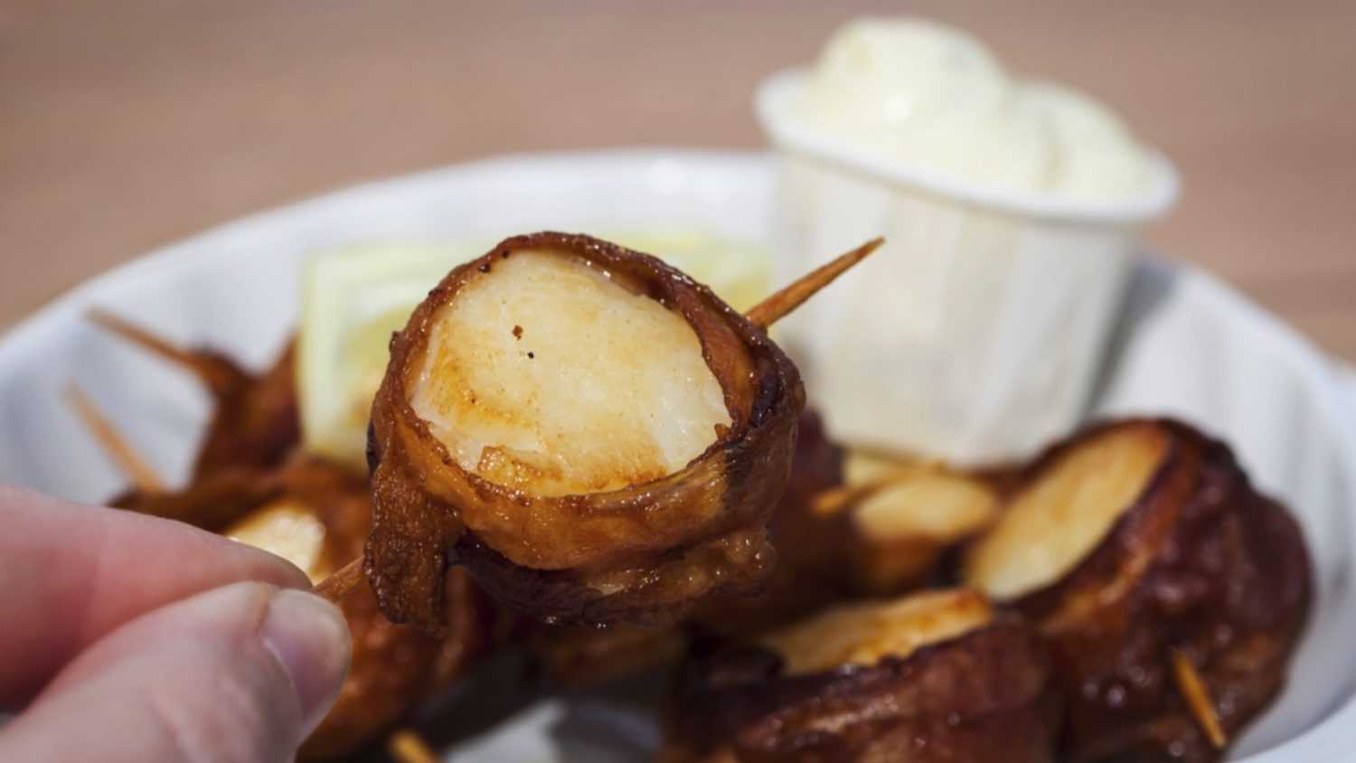 Bacon wrapped scallops in a scallop shell shaped dish at an outdoor cafe. Tarter sauce and lemon on the side.